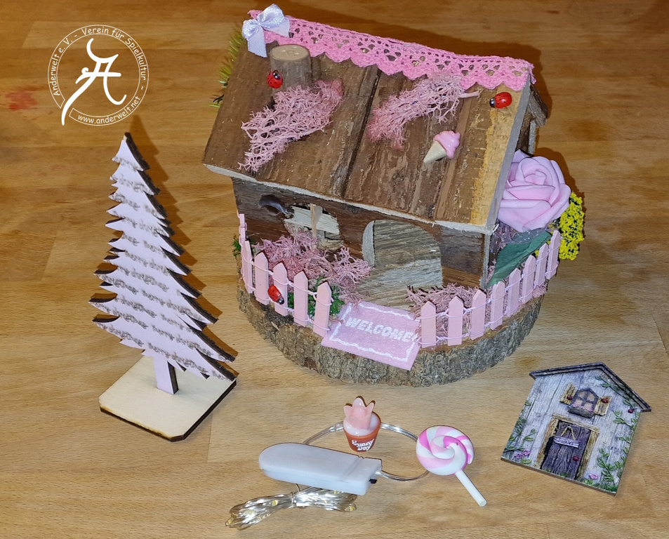 Ein kleines Holzhaus ist auf einer runden Baumscheibe aufgebaut. Ein rosa Gartenzaun umgibt es, er enthält eine Aussparung für die rosa Fußmatte mit der Aufschrift WELCOME. Der Dachgiebel ist mit einer rosa Spitzengirlande geschmückt. Eine Seitenwand des Hauses ist abnehmbar. Vor dem Haus steht eine zartrosa Tanne (Laubsägearbeit). Außerdem sind Plastik-Dekoobjekte (Süßigkeiten, Blumentopf) und eine LED-Beleuchtung zu sehen.