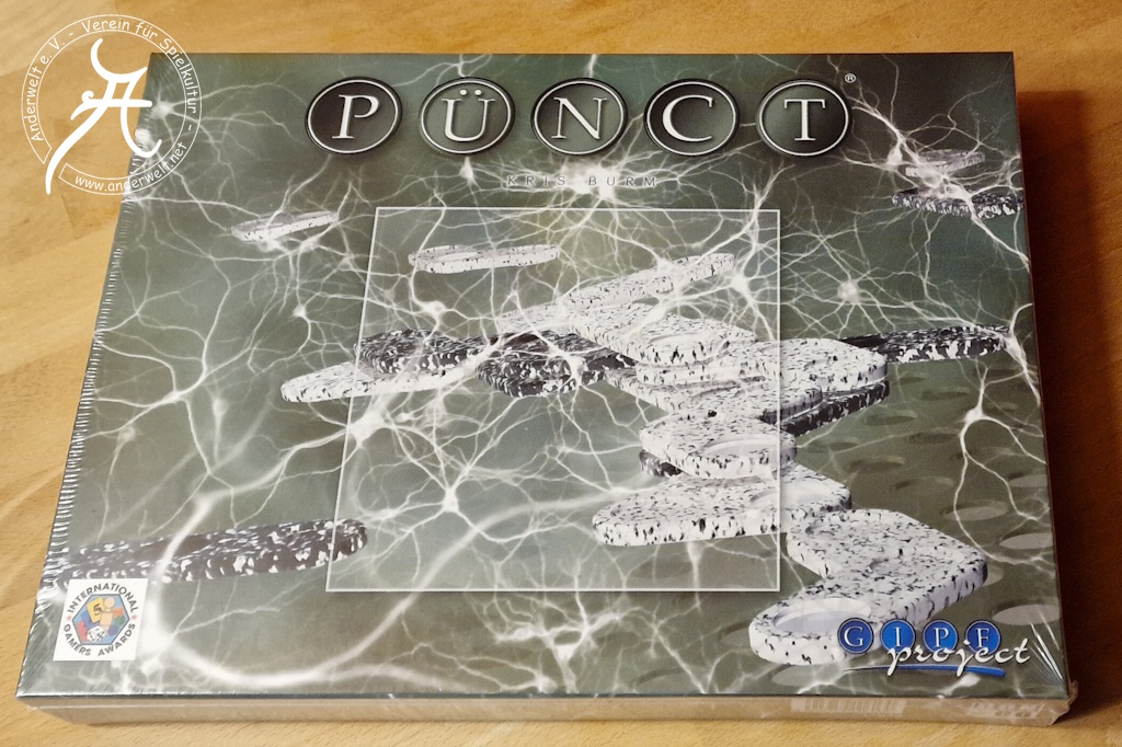 Der Shrinkwrap-verpackte Spielekarton zeigt die abgewinkelten Spielsteine in den Farben schwarz und weiß, jeweils mit einer unregelmäßigen Musterung. Sie sind umgeben von gekrümmten hellen Linien mit variabler Stärke, die an elektrische Entladungen erinnern.