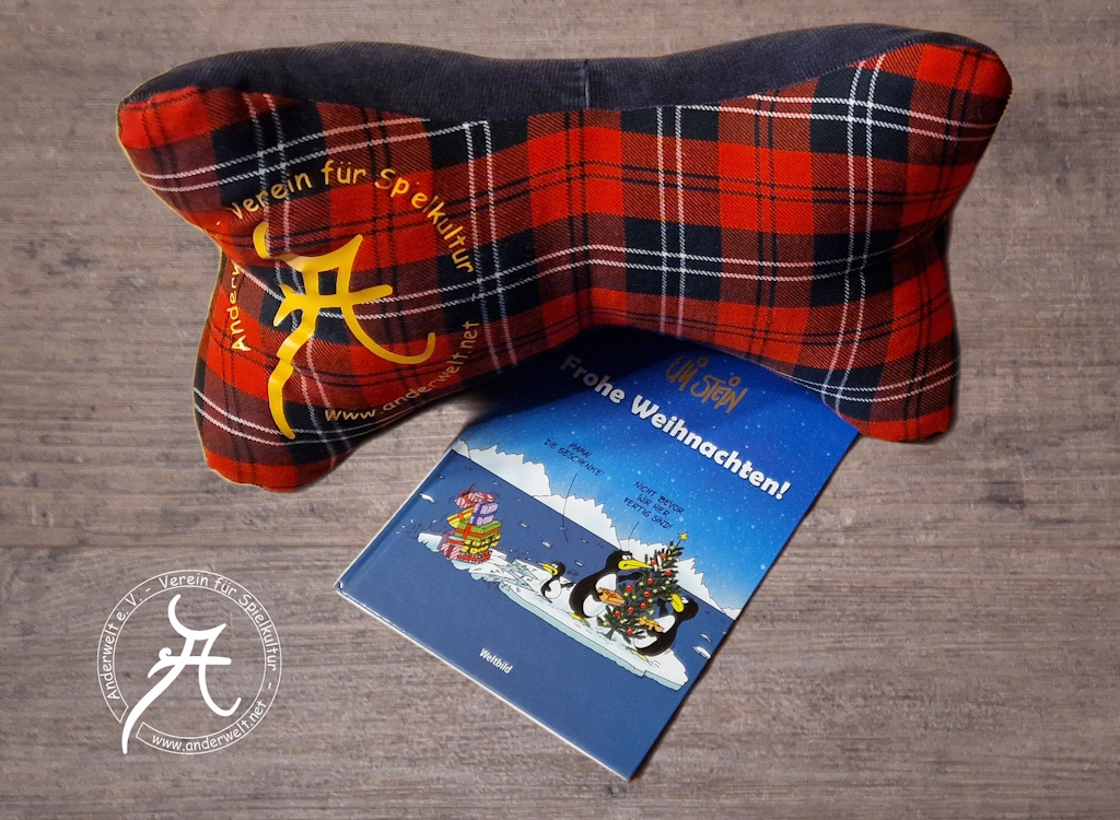 Ein Anderwelt-Leseknochen liegt auf einem Exemplar des Cartoon-Bands „Frohe Weihnachten!“ von Uli Stein. Das Titelmotiv zeigt eine Pinguin-Familie, der gerade die Weihnachtsgeschenke abhanden kommen, da die Eltern sich offenbar nicht der praktischen Risiken bewusst sind, die mit dem Klimawandel einher gehen.