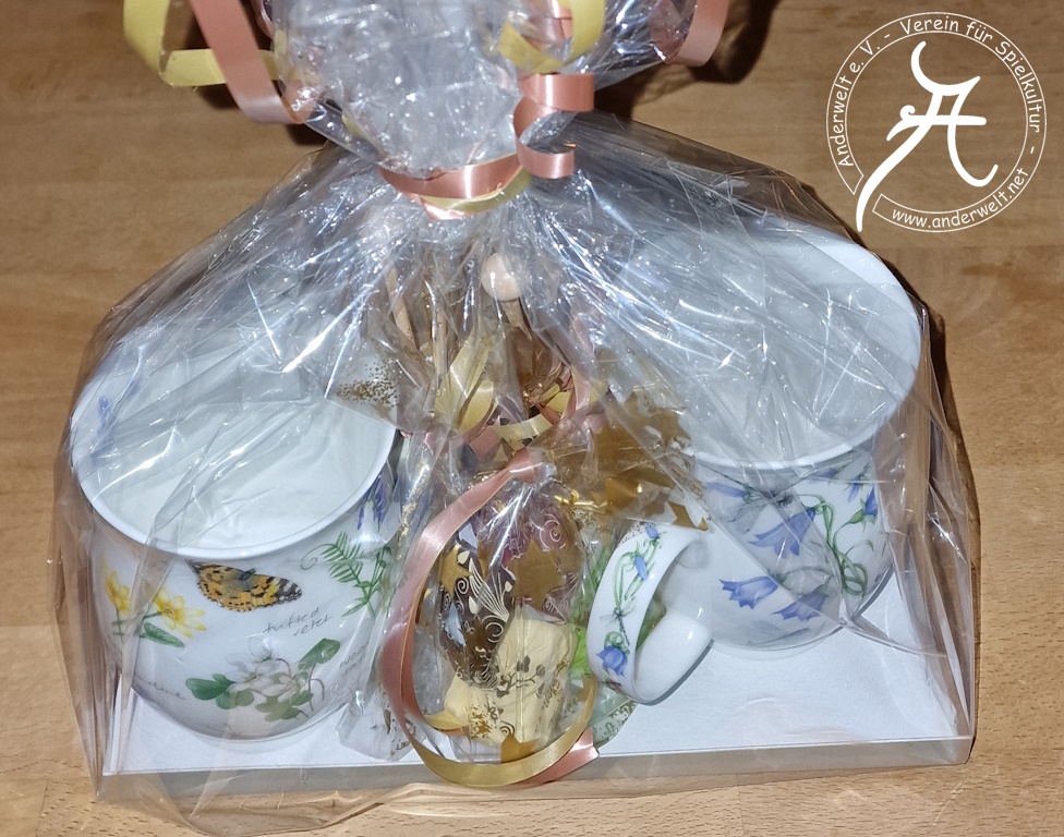 Das Bild zeigt die in Cellophan mit Geschenkband verpackten, bauchigen Teebecher mit Henkel, die von Motiven aus Flora und Fauna geziert werden. Dazwischen sind die einzeln verpackten Trüffel und Kandis-Sticks zu erkennen.