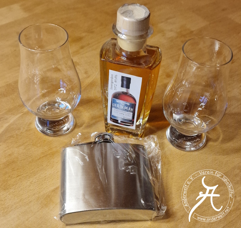 Das Bild zeigt links und rechts die beiden stiellosen Nosing-Gläser, dazwischen die verkorkte Probierflasche mit einem improvisierten Etikett, auf dem ein Foto der Originalflasche zu sehen ist. Im Vordergrund liegt der silberfarbene Flachmann, in Plastik eingeschweißt.