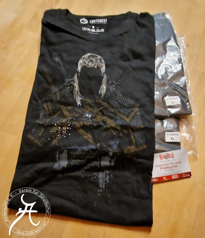 Das Bild zeigt ein ausgepacktes, aber noch weitgehend zusammengefaltetes schwarzes T-Shirt Größe M mit dem Motiv der Rüstung Geralt von Rivas sowie seinen hellgrauen Haaren. Links daneben sind die Hefte der Schwerter zu erkennen, die er auf dem Rücken trägt. Zwei weitere, noch verpackte T-Shirts in den Größen L und XL liegen darunter. Davor ist noch ein Kinopolis-Gutschein für eine Freikarte zu sehen.