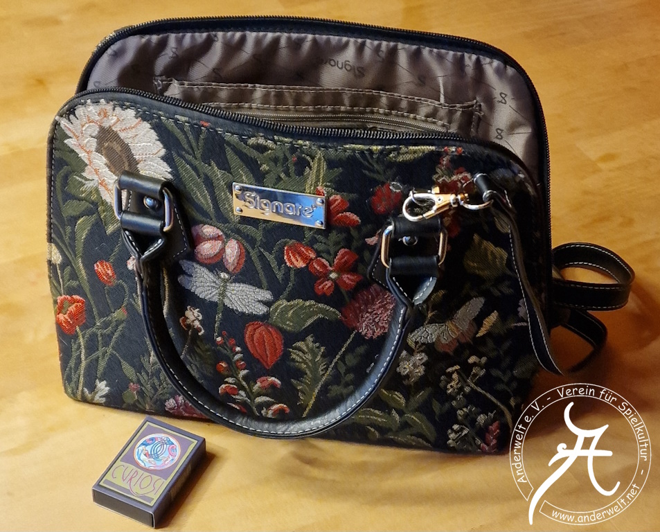 Das Bild zeigt die geöffnete Handtasche mit angebrachtem Umhängegurt. Davor liegt ein Mini-Puzzle von Curiosi im Streichholzschachtelformat.
