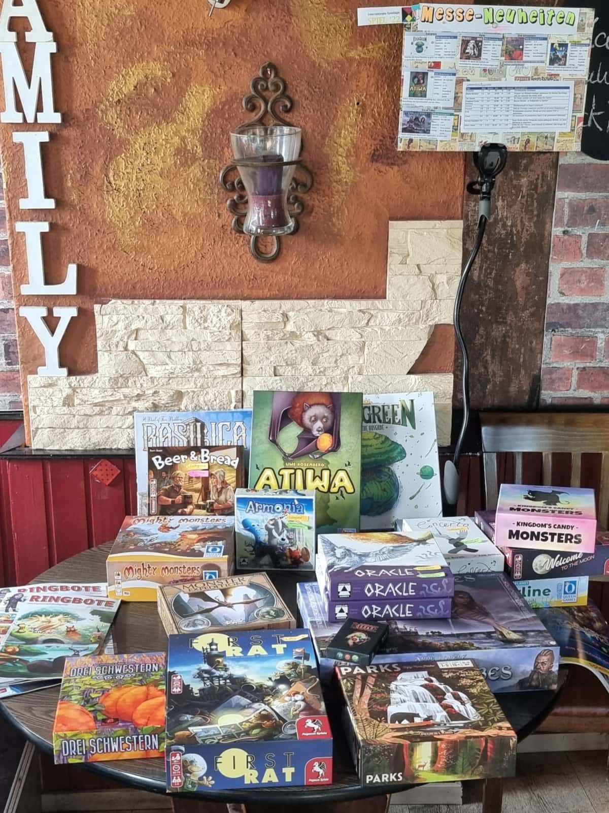 Runder Tisch, auf dem sich bunte Spiel-Schachteln stapeln. Über dem Tisch hängt ein kleines Plakat mit der Aufschrift Messe-Neuheiten.