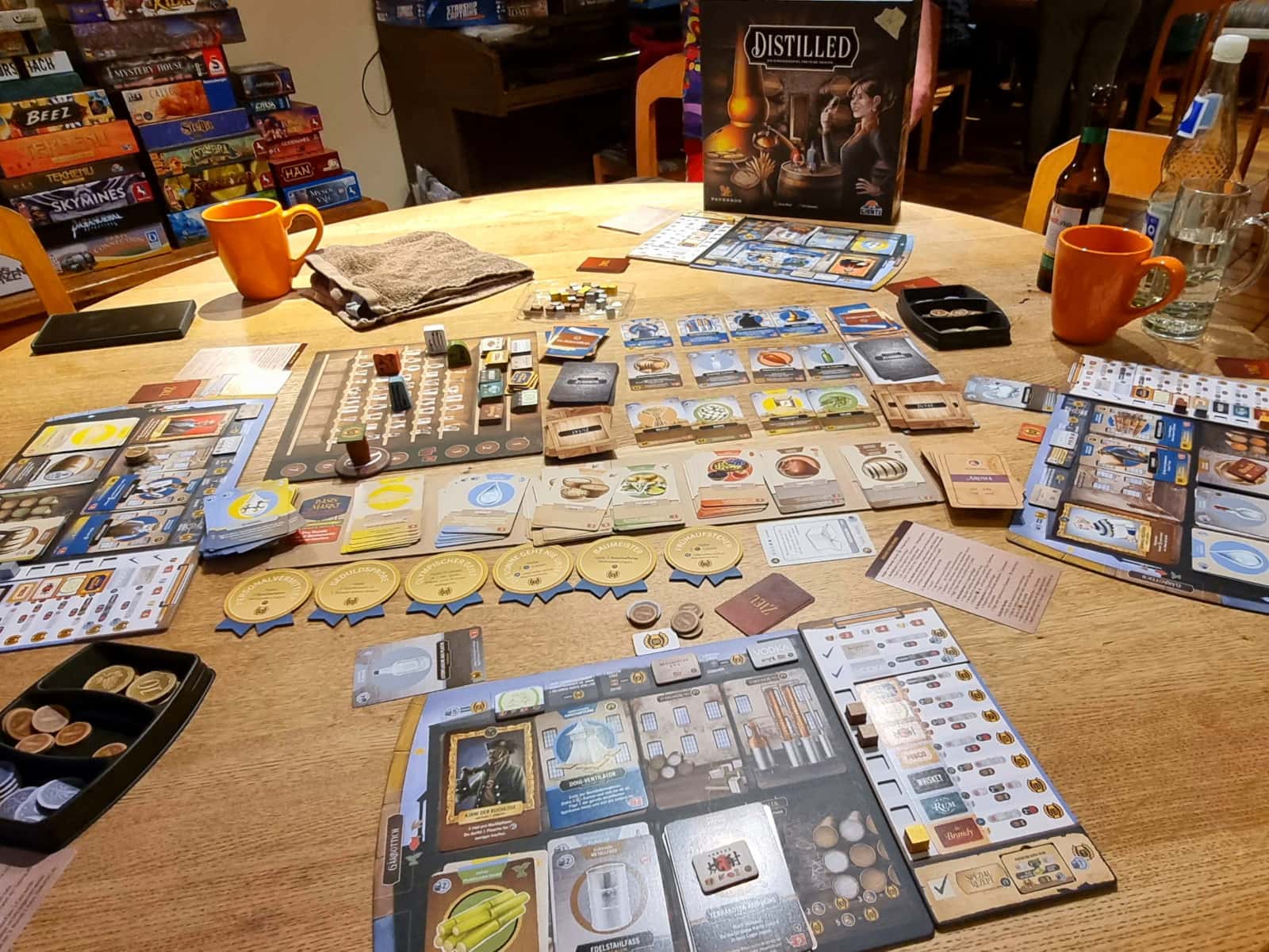 Ein ganzer Tisch voll Spielmaterial mit Spielbrett, Punkte-Brett, Markern, Marktplatzauslage, Karten und vielem mehr. Im Hintergrund ist die Spielschachtel mit einer Destille darauf zu sehen.