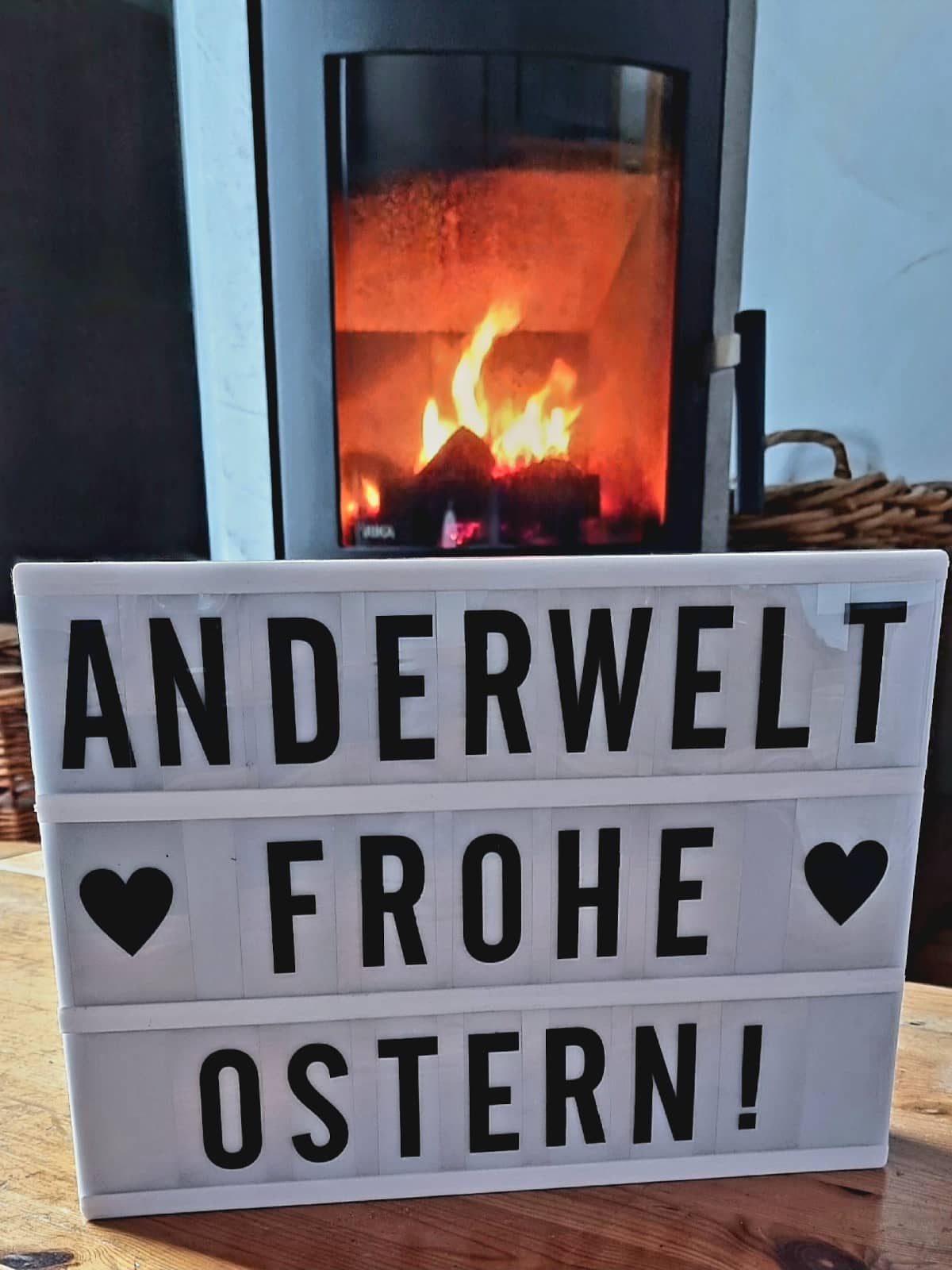 Ein Schild mit der Aufschrift "Anderwelt Frohe Ostern" vor einem loderndem Kaminfeuer.