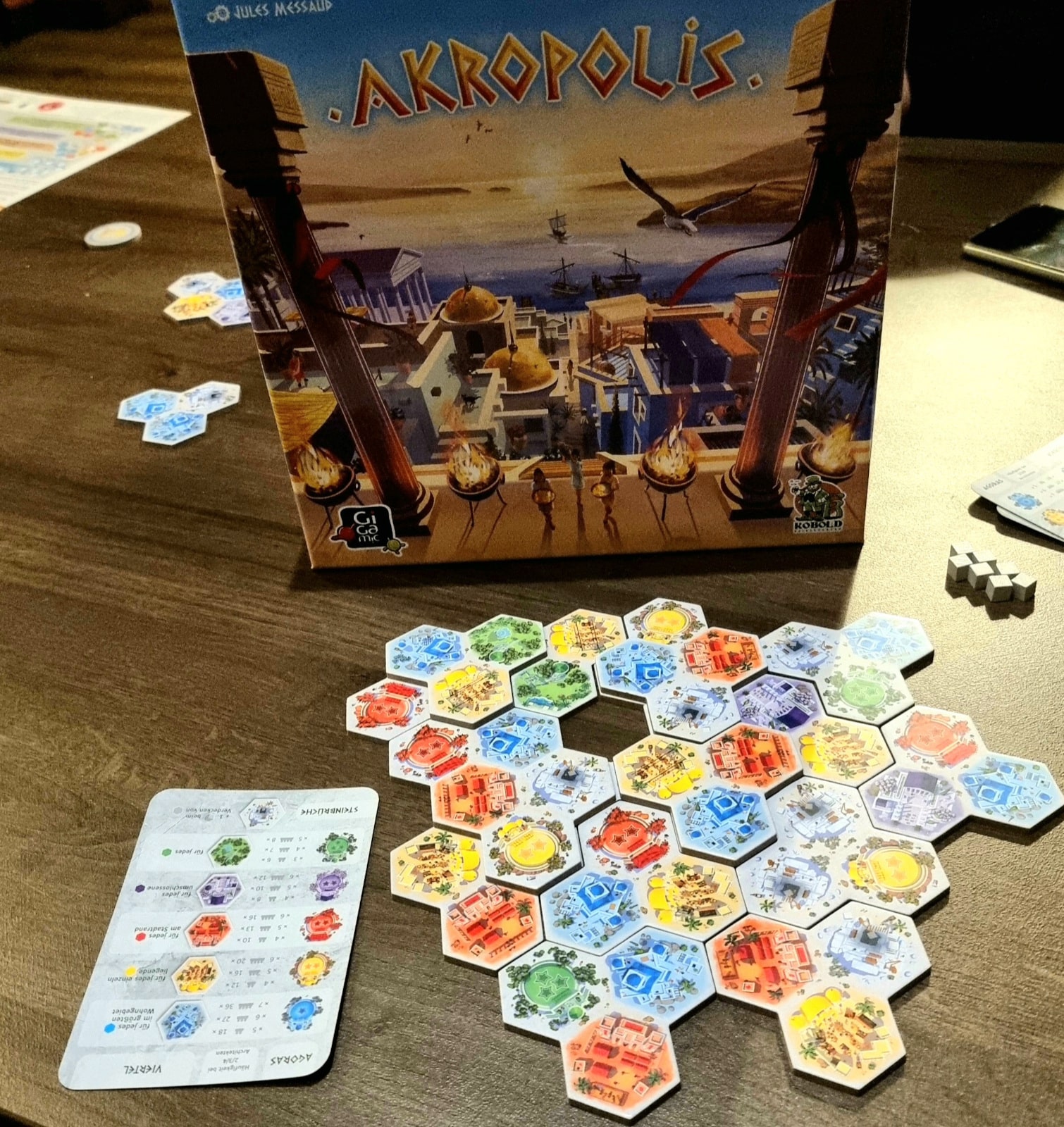 Zu sehen ist ein Tisch mit dem Spielmaterial. Im Vordergrund ist aus den Lege-Plättchen eine Stadt geformt. Einige Plättchen sind auf darunterliegende erbaut worden und bilden so terassenförmige Erhebungen. Links ist eine Hilfe-Karte mit den Lege-Regeln abgelegt. Im Hintergrund ist die aufrecht stehende Spiele-Schachtel zu sehen.