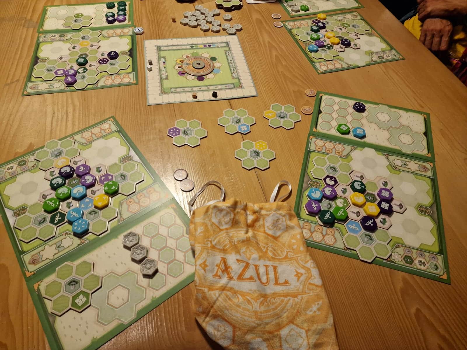 Mehrere Spieler-Pläne mit Lege-Plättchen und Spielsteinen. Frontal ist ein gelbes Azul-Säckchen zum verdeckten Nachziehen der Spielsteine zu sehen.