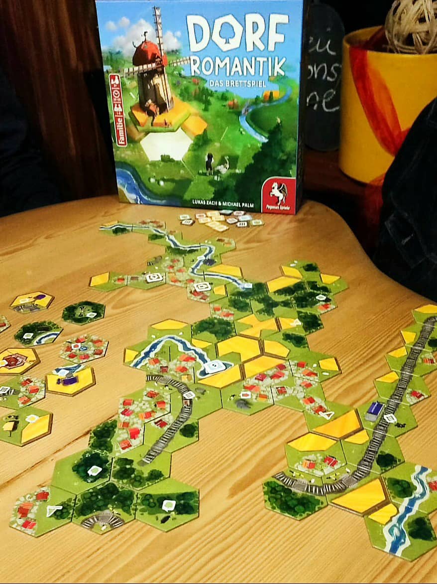 Zu sehen ist ein Tisch mit bunten sechs-eckigen Landschafts-Lege-Plättchen, die Dörfer, Felder, Flüsse, Wälder und Schienen formen. Im Hintergrund ist die aufrecht stehende Spiele-Schachtel zu sehen.