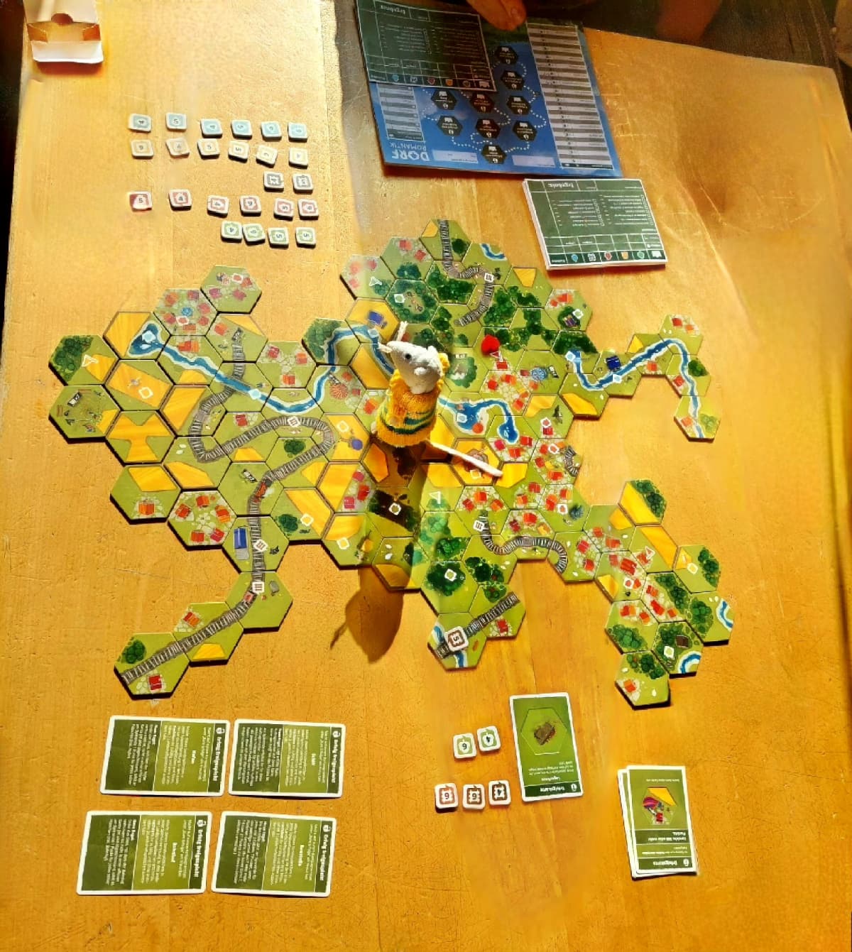 Zu sehen ist ein Tisch mit bunten sechs-eckigen Landschafts-Lege-Plättchen, die zusammenhängend Dörfer, Felder, Flüsse, Wälder und Schienen formen. Ebenfalls zu sehen sind Punkte-Plättchen, gelöste Aufgaben-Karten, ein Wertungsblock, ein Fortschritts-Block und eine kleine weiße Plüsch-Maus im Ringel-Pulover mitten auf dem Spiel-Feld.