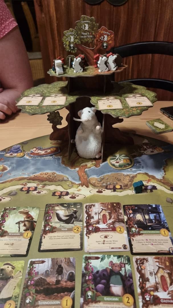 Ein Spielbrett mit Karten und großem Karton-Baum darauf. Der Baum dient als Ablagefläche für Marker. Unter dem Baum sitzt eine weiße Stoff-Maus.