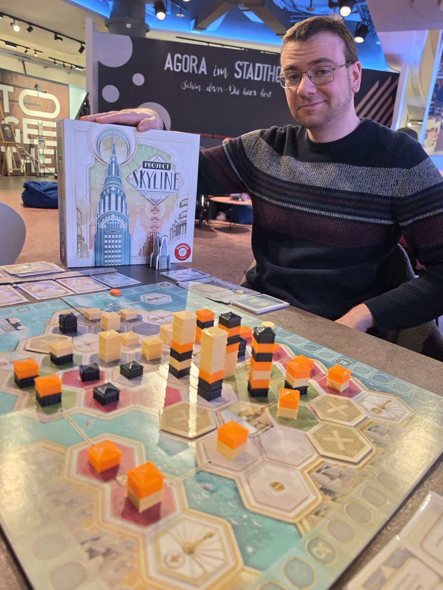 Auf einem Spieltisch ist das Spiel "Project Skyline" aufgebaut. Der Spielplan zeigt den Grundriss einer Stadt, der in hexagonale Parzellen aufgeteilt ist. Einige der Parzellen sind mit unterschiedlich hohen Gebäuden bebaut. Um den Plan herum liegen Kartenstapel. Hinter dem Tisch sitzt der Autor des Spiels, Florian Haas, und präsentiert die Schachtel.