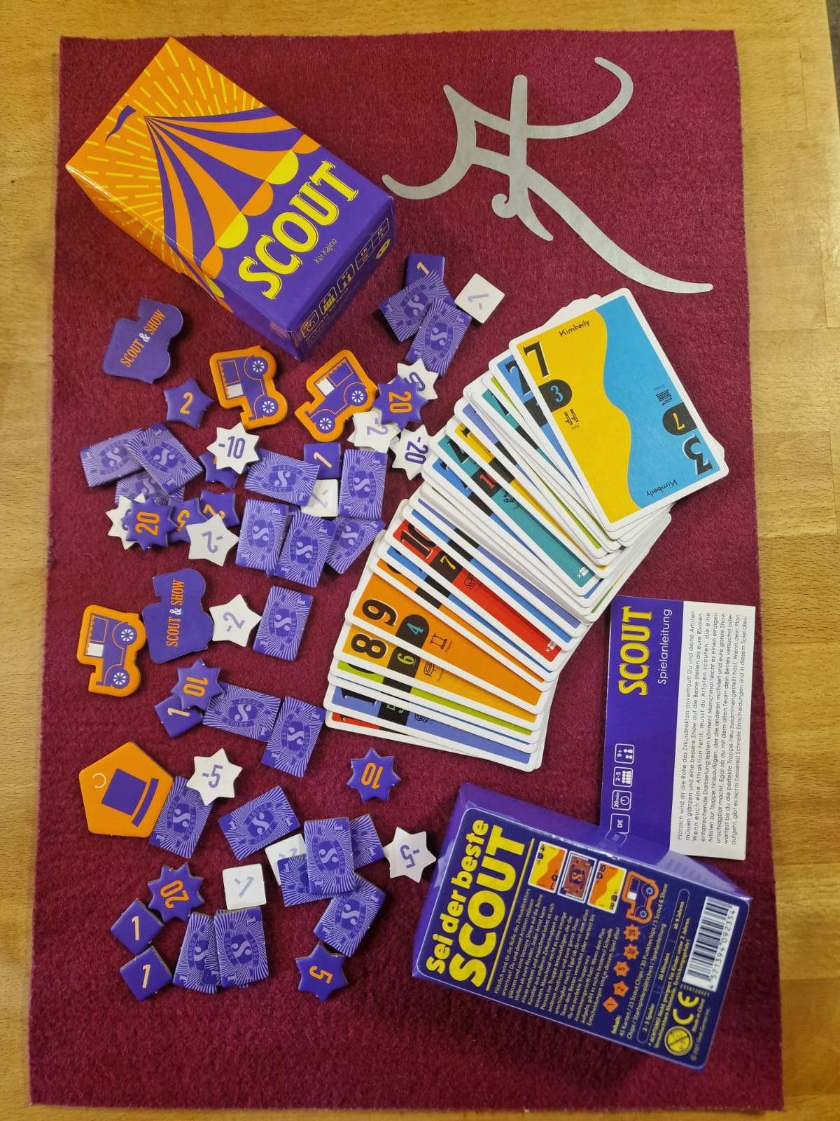 Ausgebreitetes Spielmaterial mit den zweiteiligen bunten Handkarten, den violetten Geld- und Siegpunkt-Marken, neben der minimalen Spieleschachtel.