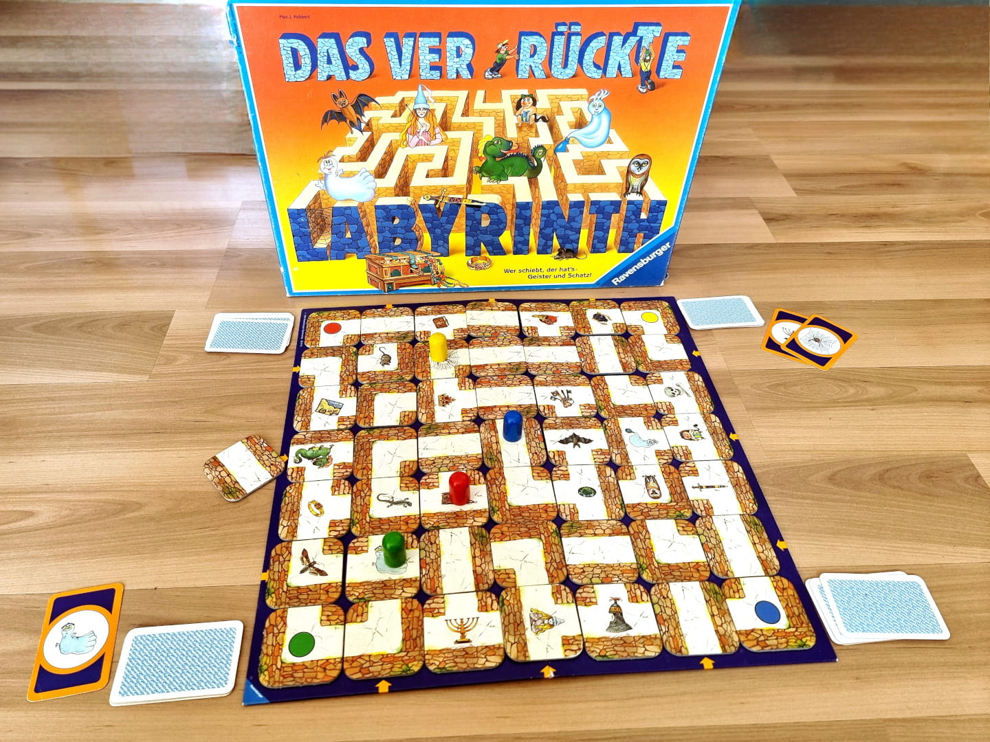 Der Spielplan aus 7x7 aneinandergelegten Weg-Plättchen bildet das Labyrinth, in dem Wesen und Objekte verstreut zu Hause sind. Am Spielrand befinden sich verdeckte als auch offene Spielkarten. Die farbigen Spielerfiguren scheinen im Spielplan eingeschlossen. Im Hintergrund ist die aufrecht stehende Spiele-Schachtel zu sehen.