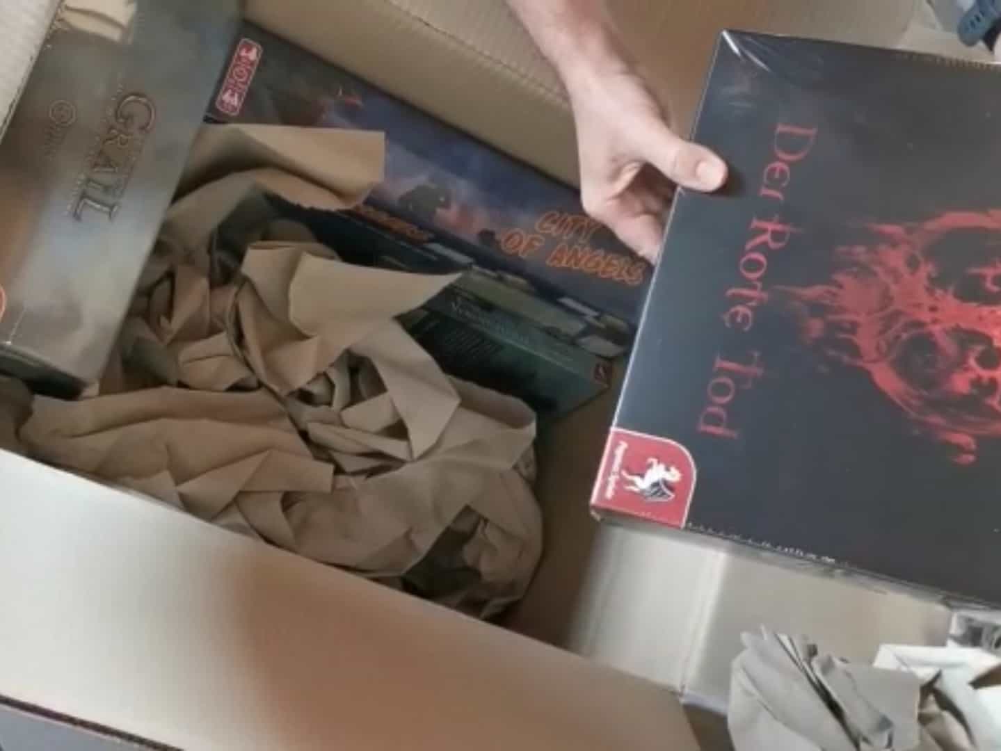 Frisch gelieferte Spiele-Kartons der Reihe Tainted Grail werden aus einem großen Karton entpackt.
