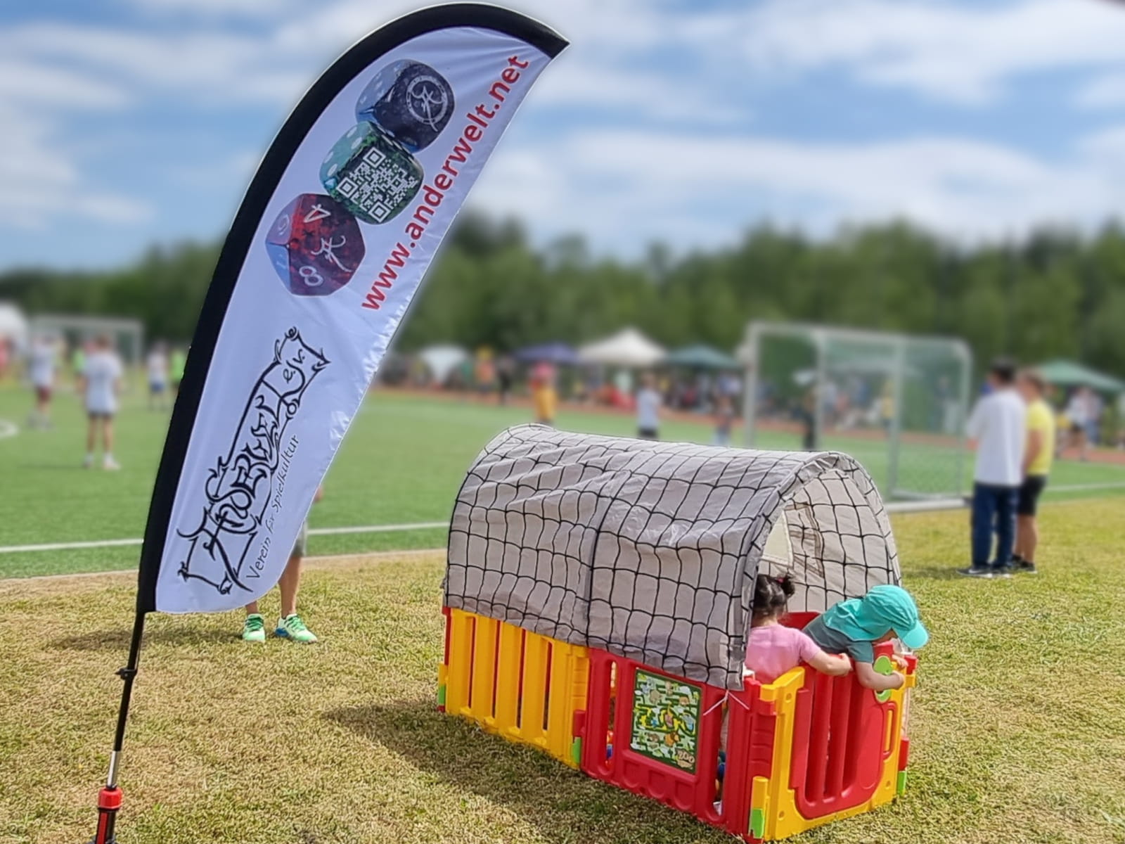 Ein mit einer Plane überspanntes Bällebad auf dem Rasen, das von Kleinkindern erkundet wird. Daneben eine eindrucksvolle Werbefahne von Anderwelt.
