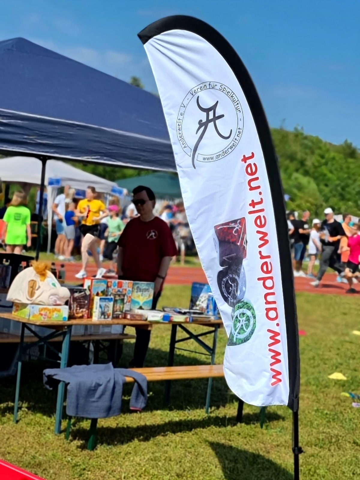 Eine Anderwelt-Werbe-Fahne mit verschiedenen Anderwelt-Würfeln, die vor einem Zelt-Stand aufgestellt ist. Am Stand werden verschiedene Brettspiele präsentiert.