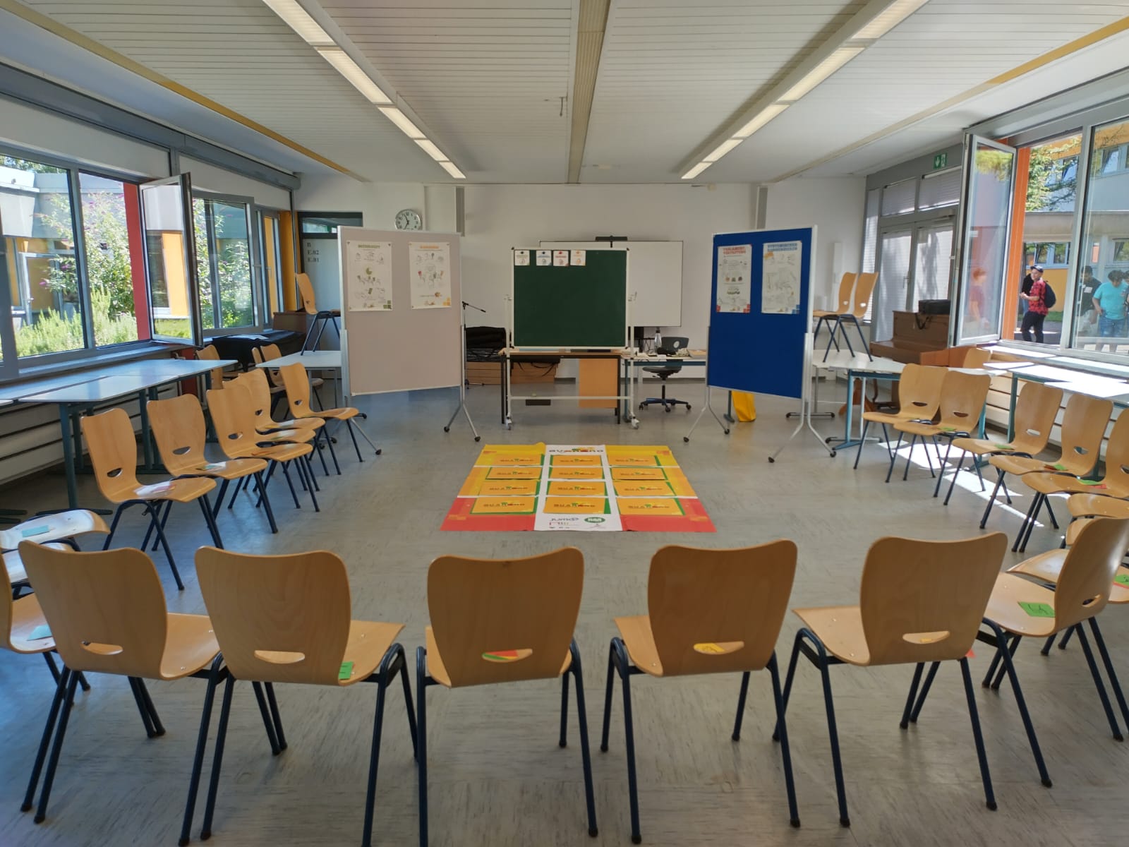 Zu sehen ist ein heller Raum mit einem Stuhlkreis und verschiedensten Lehrmaterialien sowie einer Pinwand.