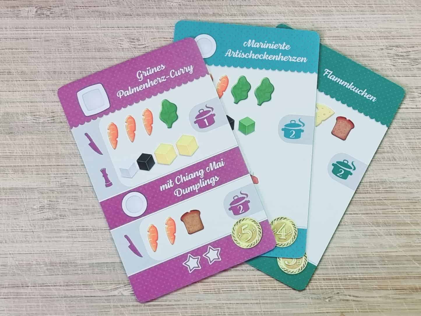 Drei Rezept-Karten mit erforderlichen Zutaten von einem Brettspiel liegen auf einem Holzbrett.