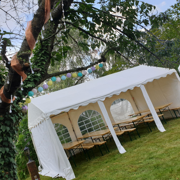 Ein Festzelt mit Tischen und Bänken im grünen Garten. Über dem Zelt bäumen sich Äste eines großen Baumes, die mit Lichterketten und bunten Lampions geschmückt sind.