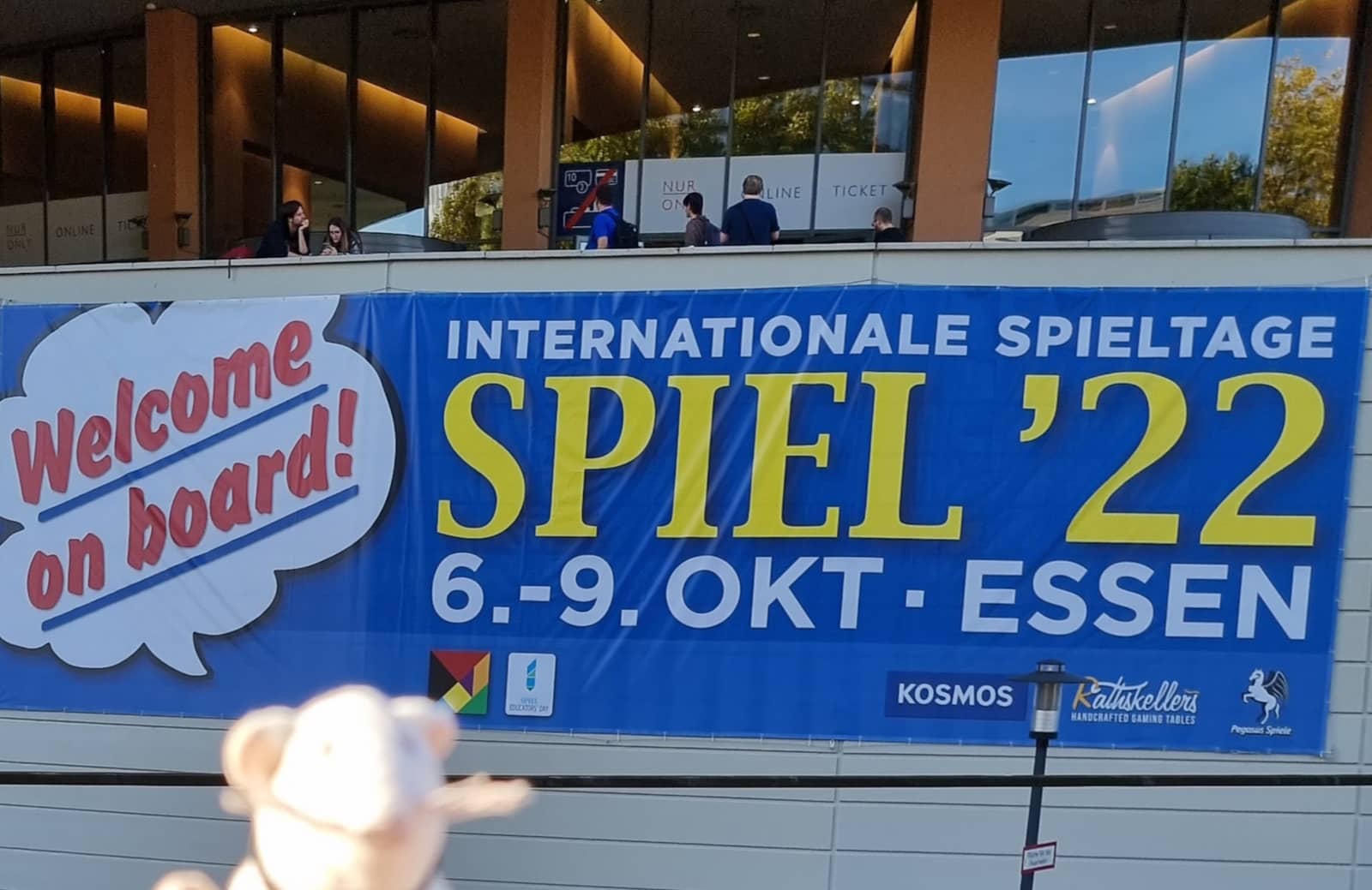 Zu sehen ist der Messe-Eingang der SPIEL 2022 in Essen. Davor ein großes  blaues Werbebanner für die Messe mit der bekannten Sprechblase: 
