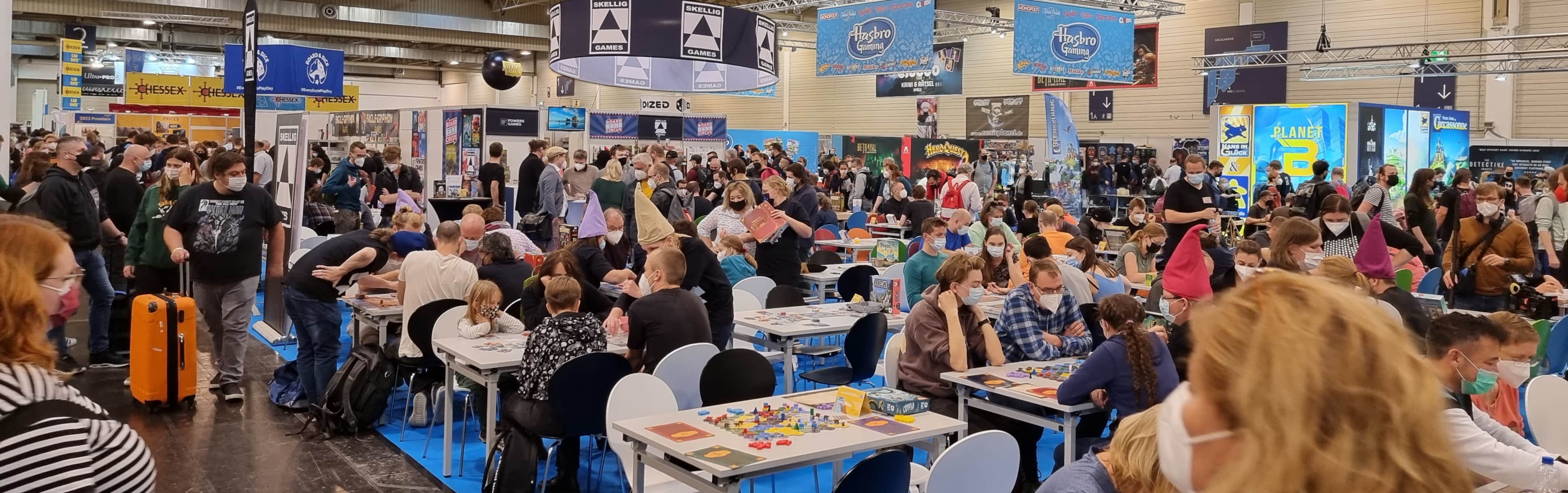 Viele besetzte Spieltische mit Messe-Trubel drumherum. Im Hintergrund sind die Schilder und bunten Werbe-Plakate der Verlage zu sehen.
