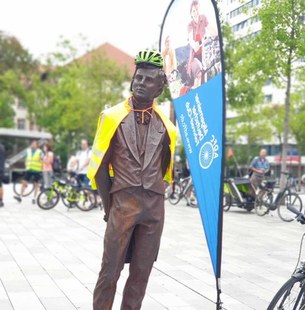 Eine mit Fahrradhelm und Warnweste ausgerüstete Statue am Hanauer Freiheitsplatz, daneben eine Werbefahne vom ADFC. 