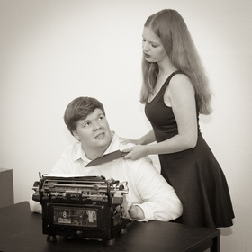 Eine Frau hällt einem Mann, der verängstigt vor einer altmodischen Schriebmaschiene sitzt, ein großes Küchen-Messer an die Kehle.