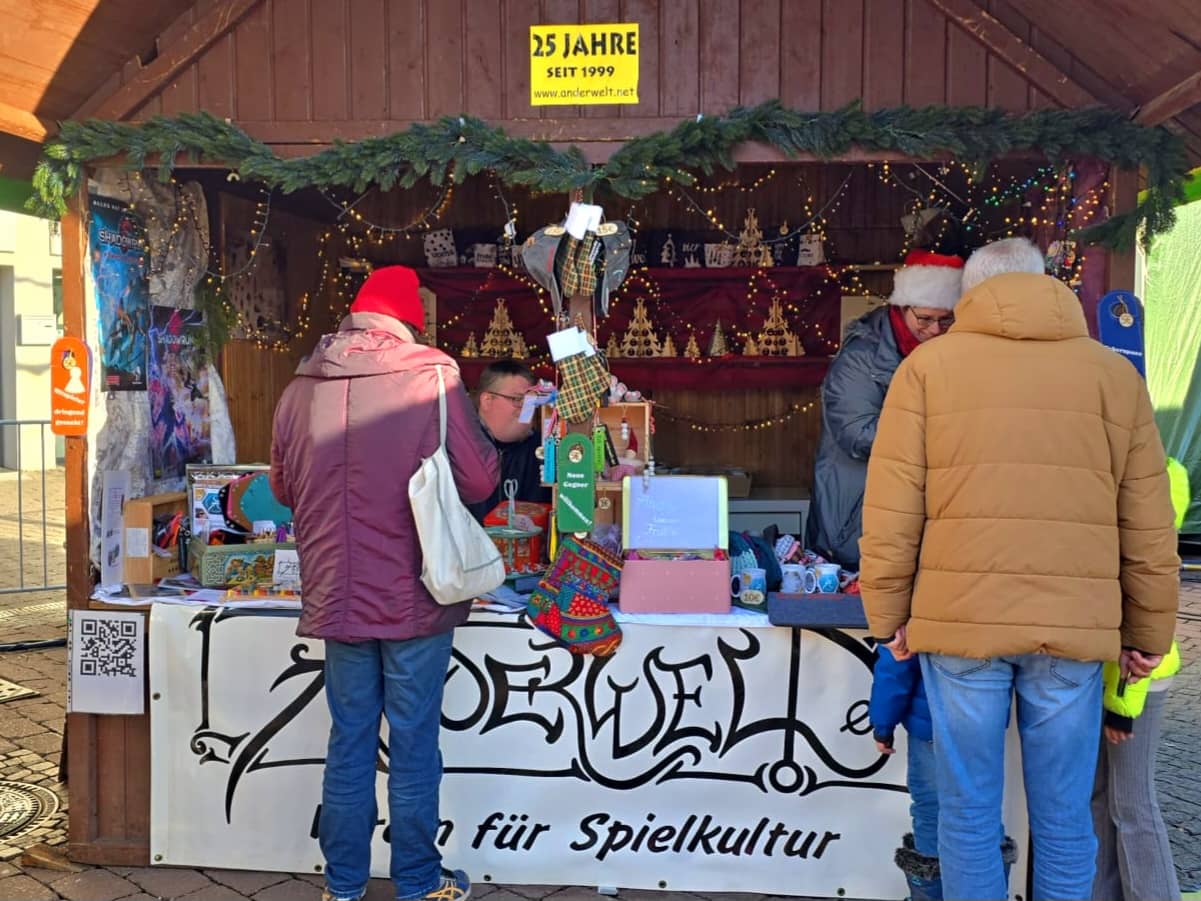Weihnachtlich geschmückter Verkaufsstand auf dem Markt mit Anderwelt-Transparent, Verkaufsware, Helfern und Kunden.