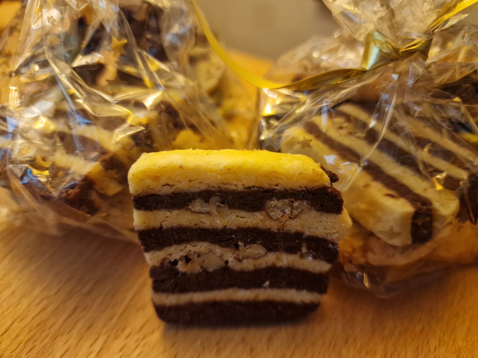 Ein aufrecht stehendes, rechteckiges Plätzchen, das in schmalen Streifen von dunklem und hellem Teig durchzogen ist. Im Hintergrund sind abgepackte Geschenk-Tütchen mit diesen Plätzchen zu sehen.
