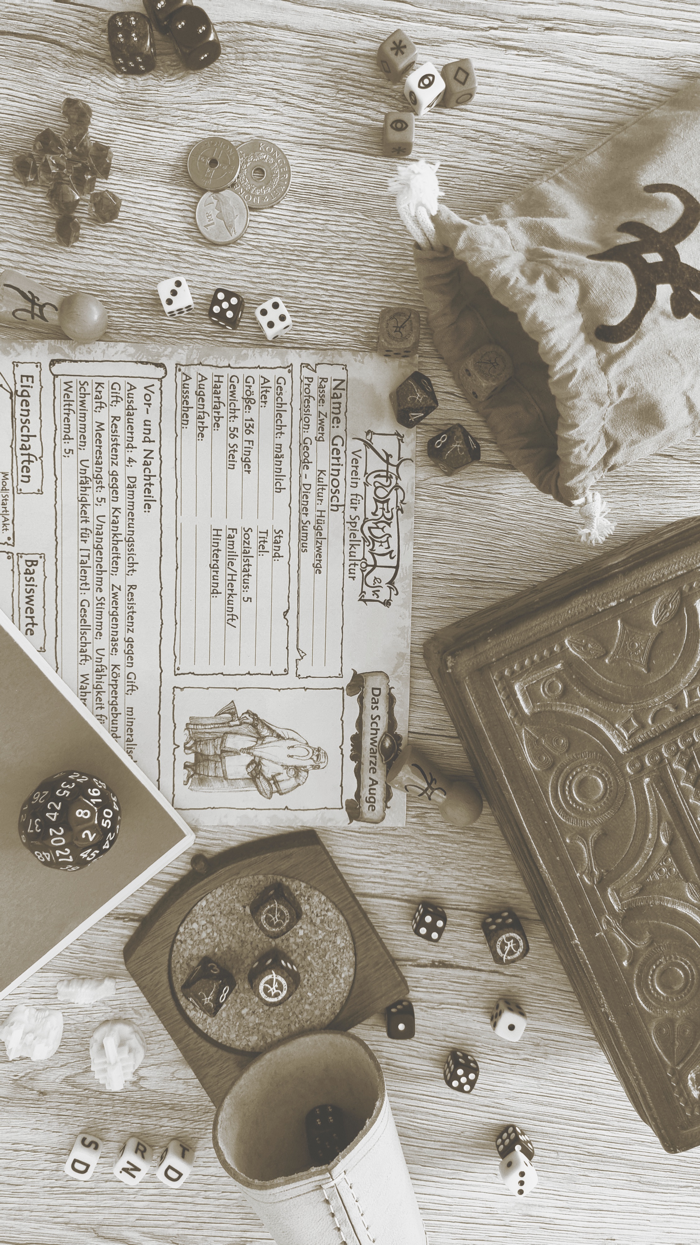 Viele Rollenspiel-Utensilien auf einem Tisch verteilt. Darunter diverse Würfel (W6,W8,W50), Münzen, Edelsteine, ein Charakter-Bogen eines Zwerges von DSA, eine Schatulle, ein Anderwelt-Würfelbeutel, ein Schreib-Heft und ein Würfelbecher mit Untersetzer.