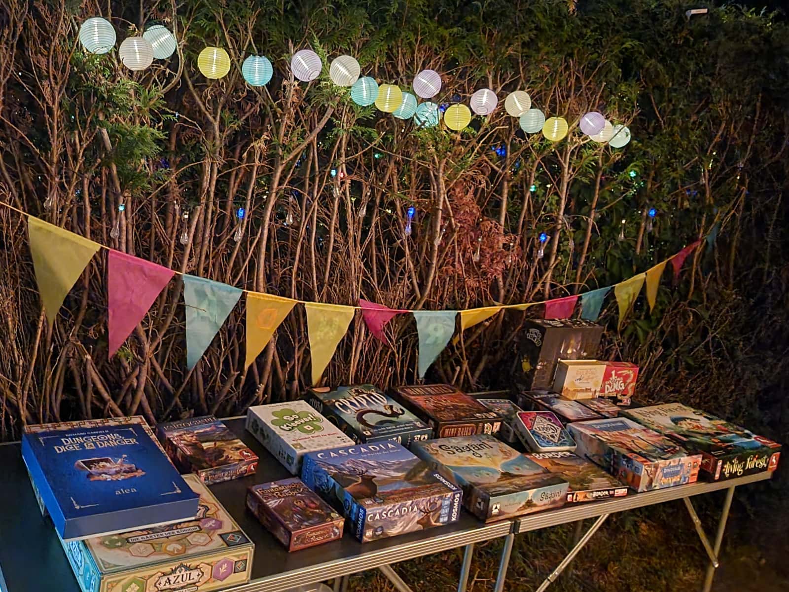 Aufgebahrtes Spiele-Angebot auf einem langen Tisch vor einer Hecke, dahinter bunte Lampions, Wimpel und Lichterketten.